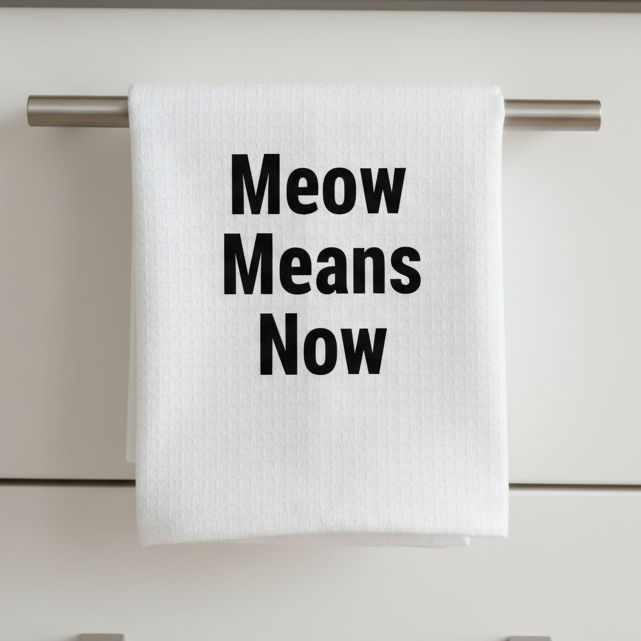 white kitchen tea towel with the words "Meow Means Now" in black font on the towel. The towel is hanging from a stainless steel handle